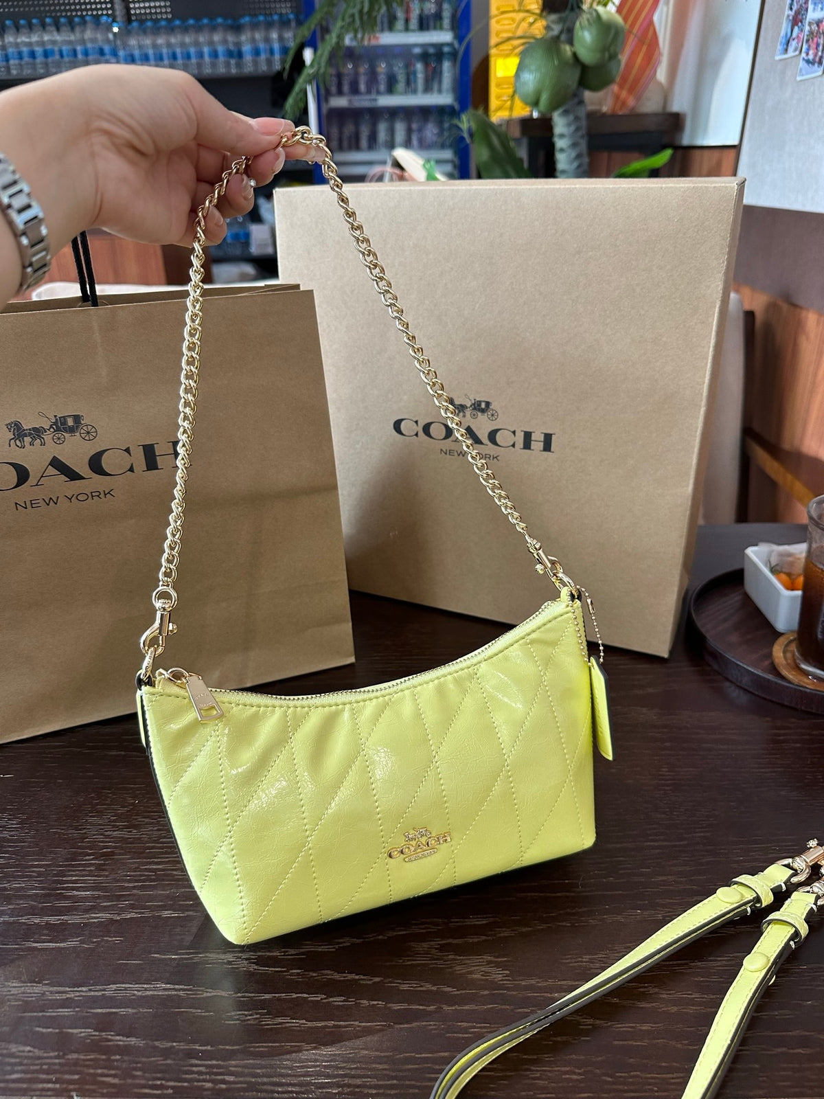 A Coach shoulder bag in light yellow patent leather with a gold chain strap, held by a hand in front of its brown Coach shopping bags on a dark wooden table.