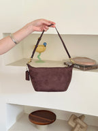 A person's hand holds up a dark brown suede shoulder bag with gold-tone hardware and a matching leather tassel, set against a minimalist white shelving background.