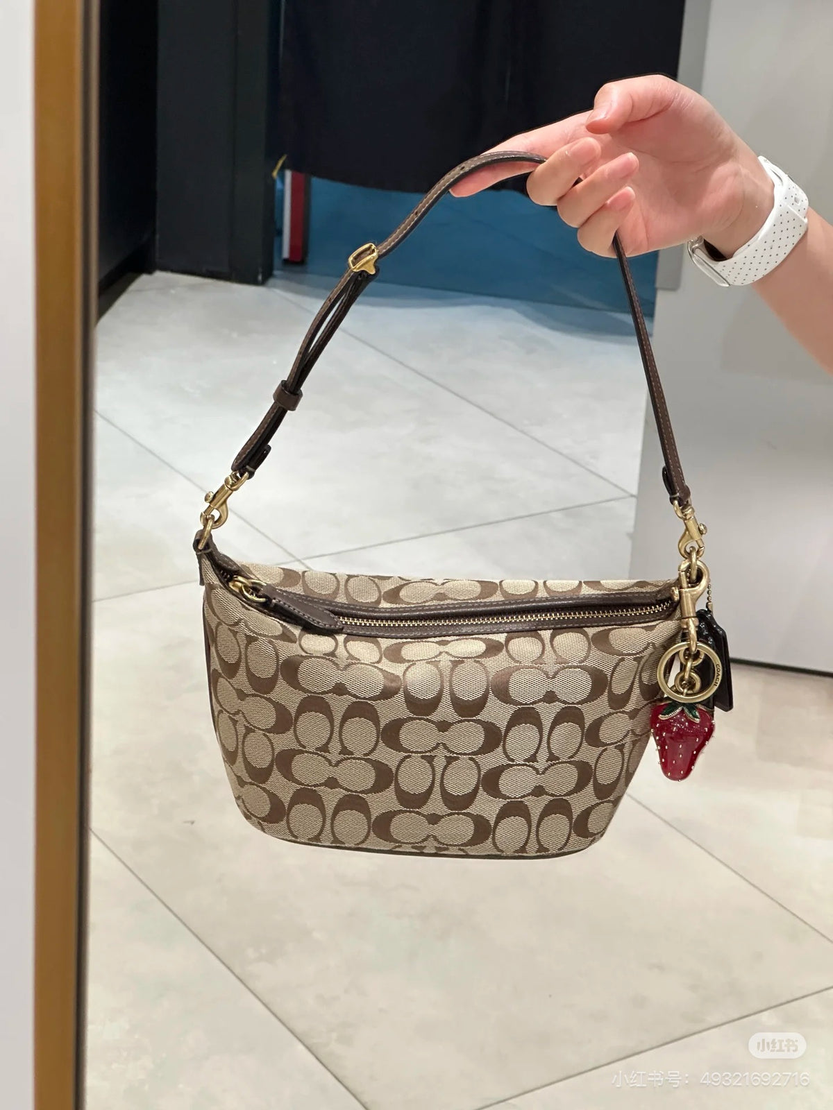 A Coach signature monogram shoulder bag in beige and brown canvas with gold-tone hardware, held by a person's hand in a store setting.