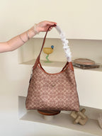 A person's arm holds up a Coach hobo bag with a brown and beige monogram pattern. The bag features a white ruffled fabric sleeve wrapping around the shoulder strap, set against a minimalist beige wall with built-in shelves containing decorative items.