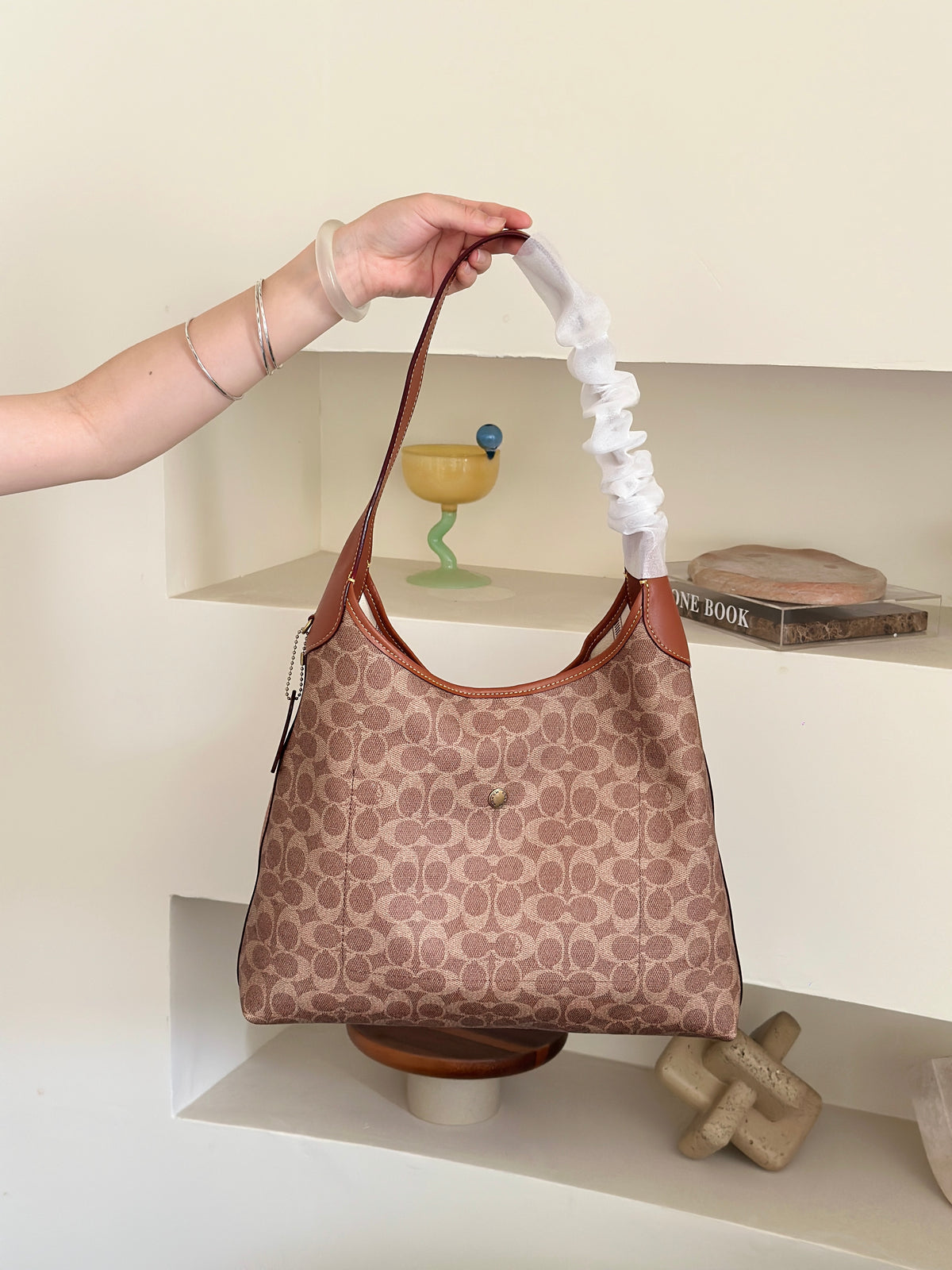 A person's arm holds up a Coach hobo bag with a brown and beige monogram pattern. The bag features a white ruffled fabric sleeve wrapping around the shoulder strap, set against a minimalist beige wall with built-in shelves containing decorative items.