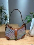 A multicolor patterned canvas shoulder bag with brown leather trim and strap, displayed on a wooden table with a cardboard box and green plants in the background.