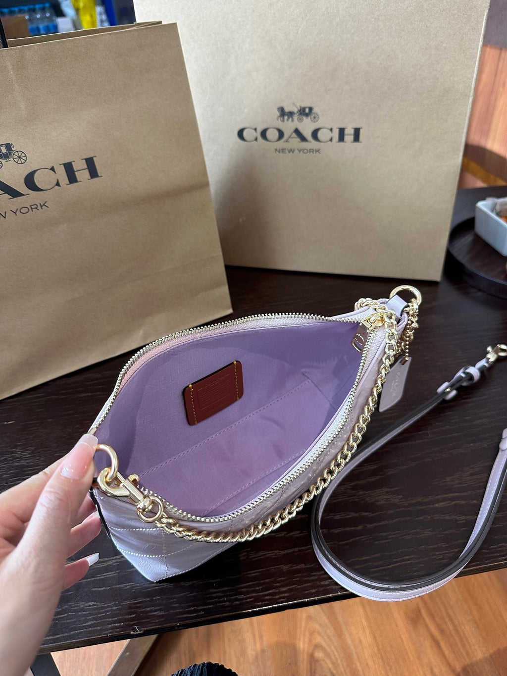 A Coach shoulder bag in light lavender purple nylon with gold-tone chain and zipper hardware, held open by a hand. The bag features a lavender interior, a small brown leather logo patch, and a white leather strap.