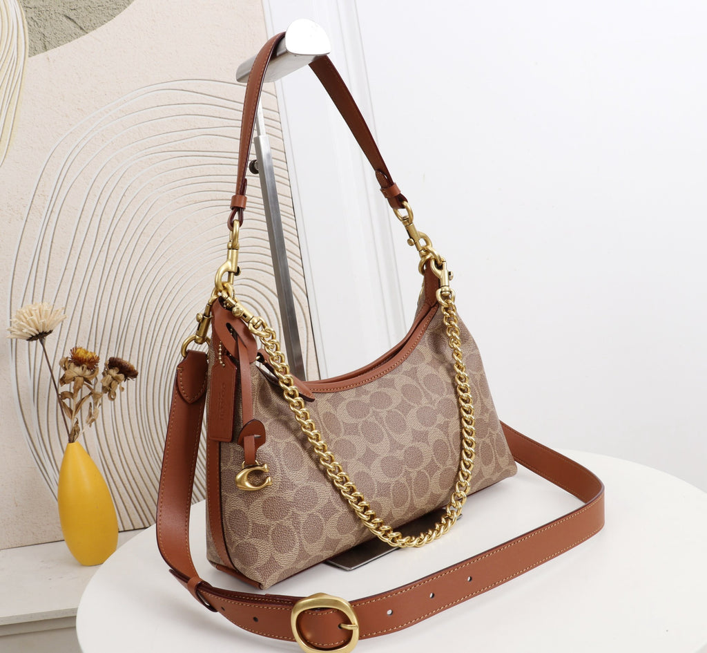 A Coach hobo shoulder bag in the signature monogram canvas, featuring brown leather trim, a gold-tone chain strap, and a matching brown leather shoulder strap, displayed on a white table with a decorative background.