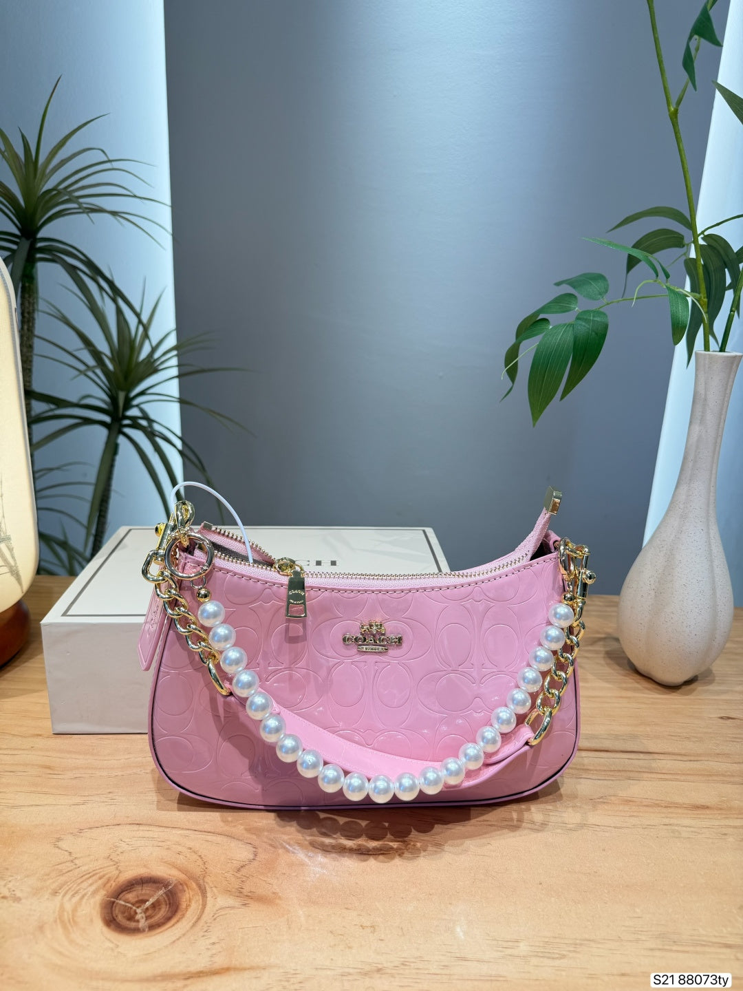 A pink embossed leather shoulder bag with a pearl strap and gold-tone chain accents, displayed on a wooden table with a white box and indoor plants in the background.