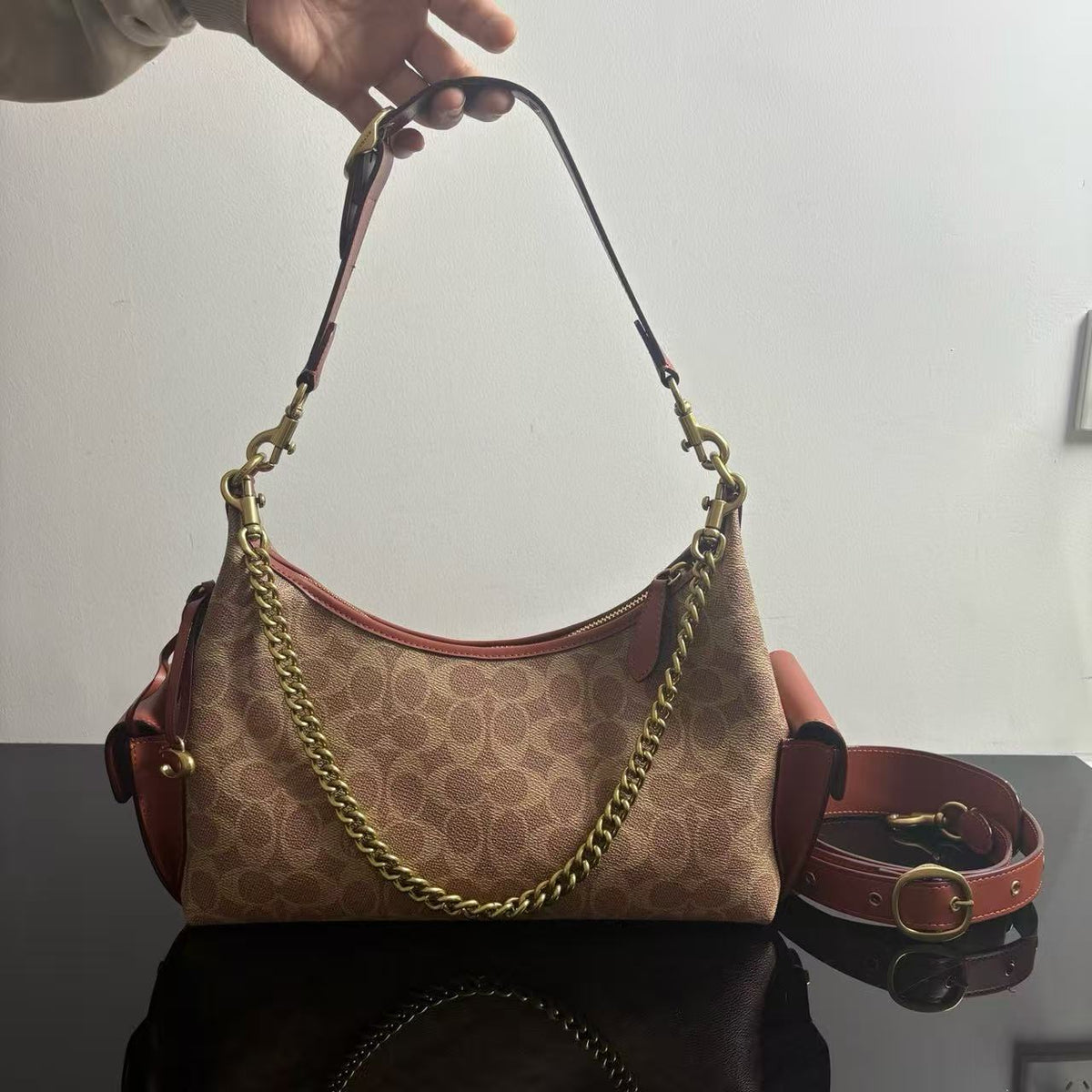 A Coach hobo shoulder bag in the signature monogram canvas, held by a hand. The bag features brown leather trim, a gold-tone chain strap, and a matching brown leather shoulder strap resting on a dark reflective surface.