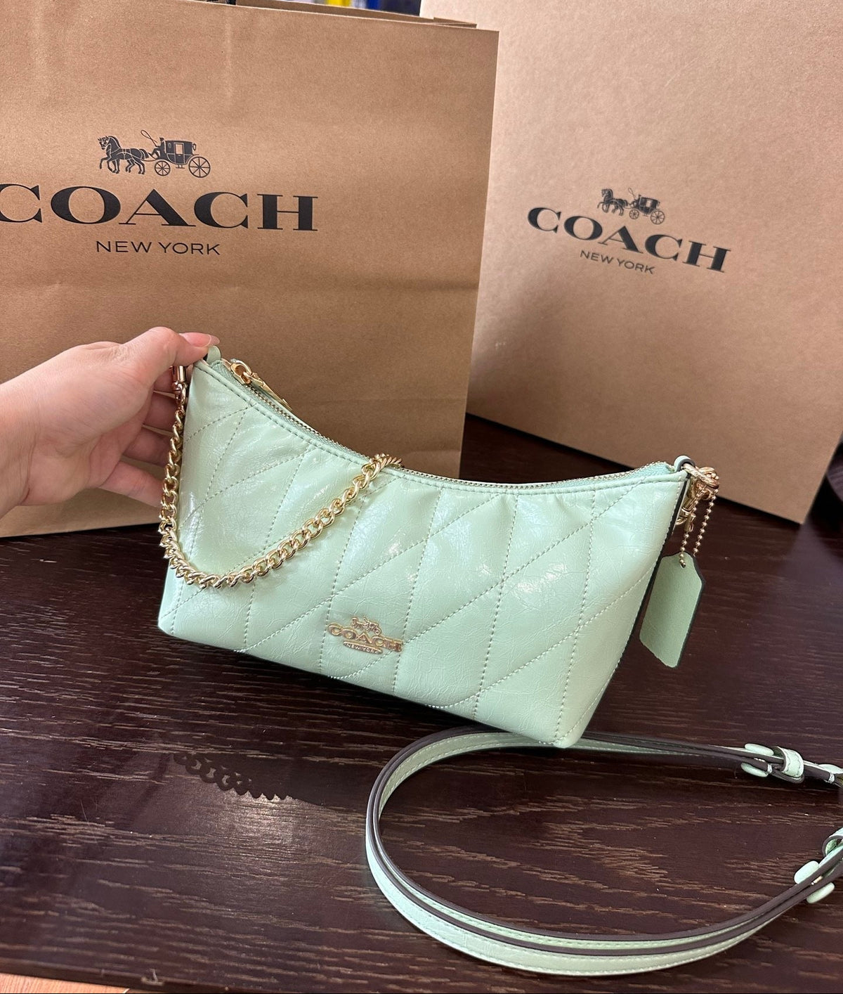 A light mint green Coach quilted shoulder bag with gold hardware is held by a hand in front of two brown Coach New York shopping bags on a dark wooden table.