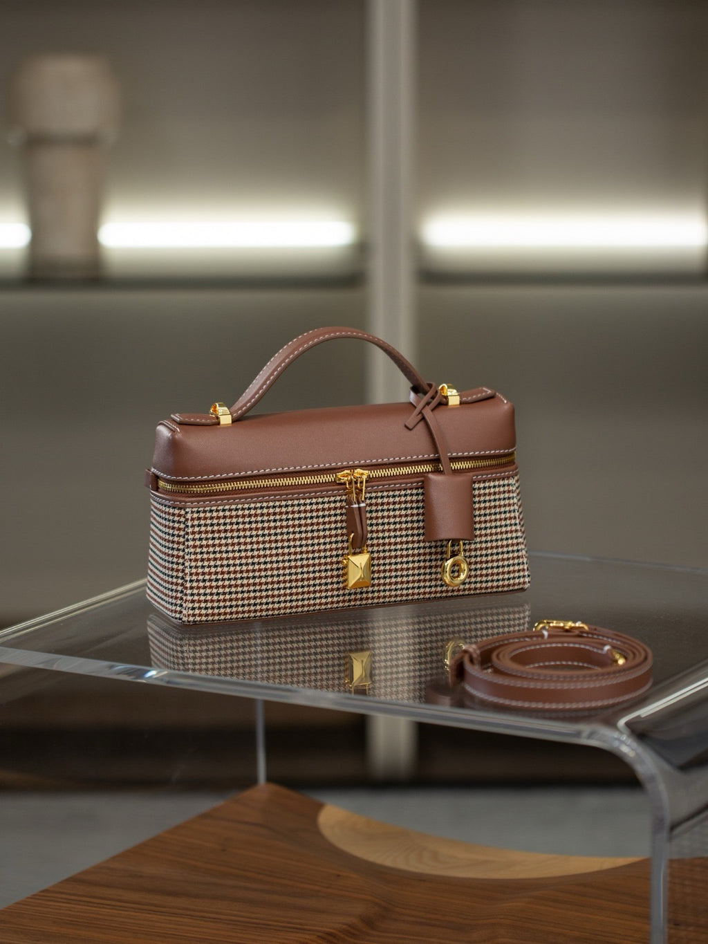A structured handbag with a brown leather top handle and trim, combined with a beige and brown houndstooth patterned body, displayed on a glass table with its matching brown strap nearby.