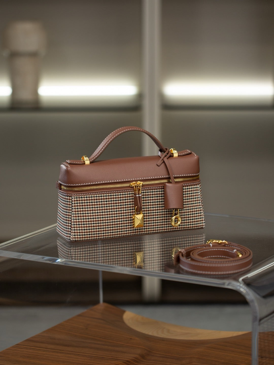 A structured handbag with a brown leather top handle and trim, combined with a beige and brown houndstooth patterned body, displayed on a glass table with its matching brown strap nearby.