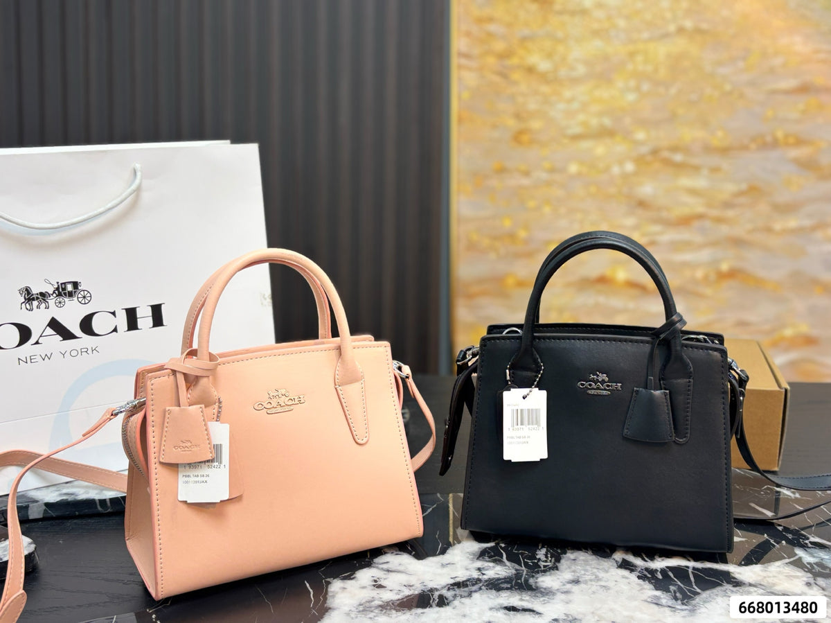 Two Coach handbags displayed side by side on a marble surface, with a Coach shopping bag and box in the background. The left bag is a light peach-pink leather top-handle satchel, and the right bag is a black leather top-handle satchel.