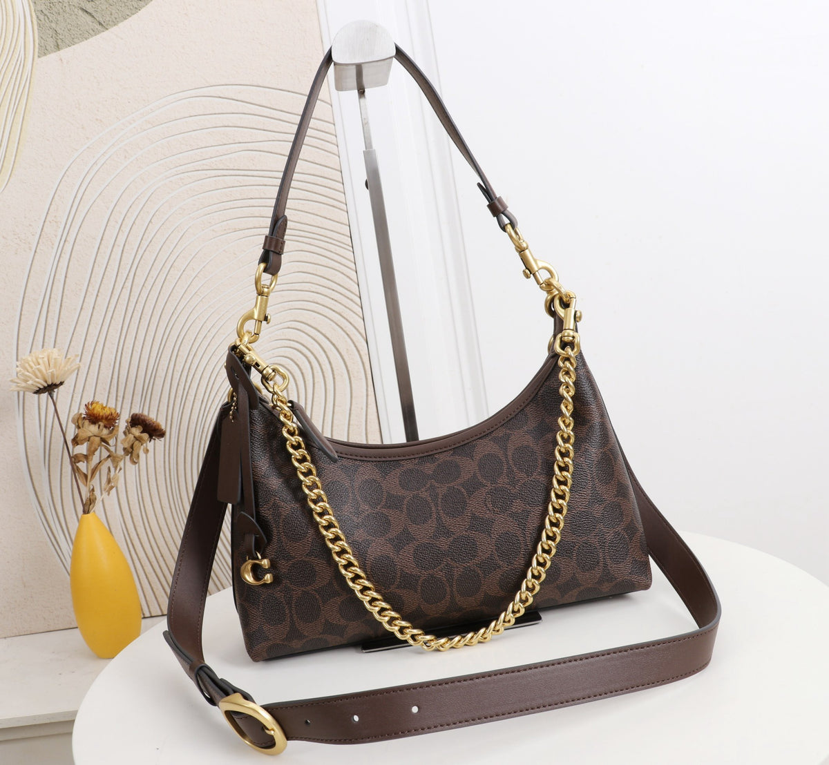 A brown monogram shoulder bag with gold-tone chain and hardware, displayed on a white table against a light background with a decorative vase.
