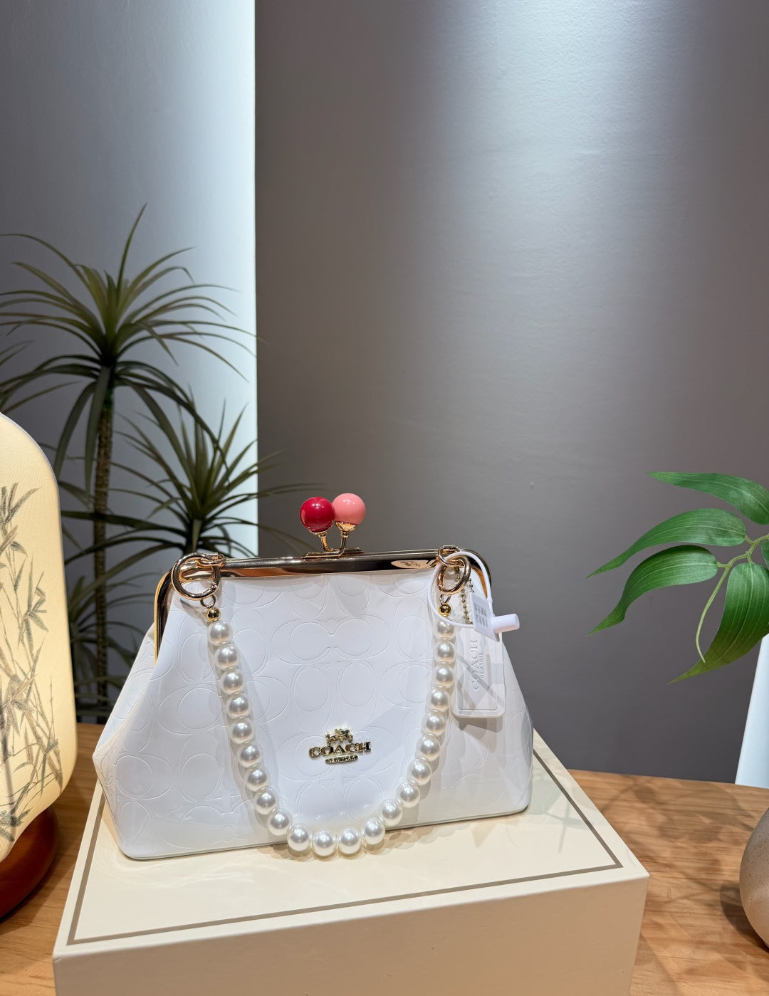 A white Coach handbag with a pearl-look strap and a gold-tone kiss-lock closure, displayed on a cream-colored box in an indoor setting with plants in the background.