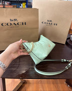 A mint green Coach quilted shoulder bag held by a hand, with two brown Coach shopping bags in the background on a dark wooden table.