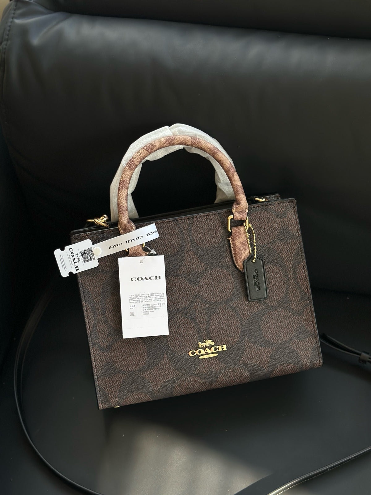 A Coach mini top-handle bag in brown signature monogram canvas, resting on a black leather chair. The bag features a brown and beige patterned handle wrap, gold-tone hardware, and white product tags.