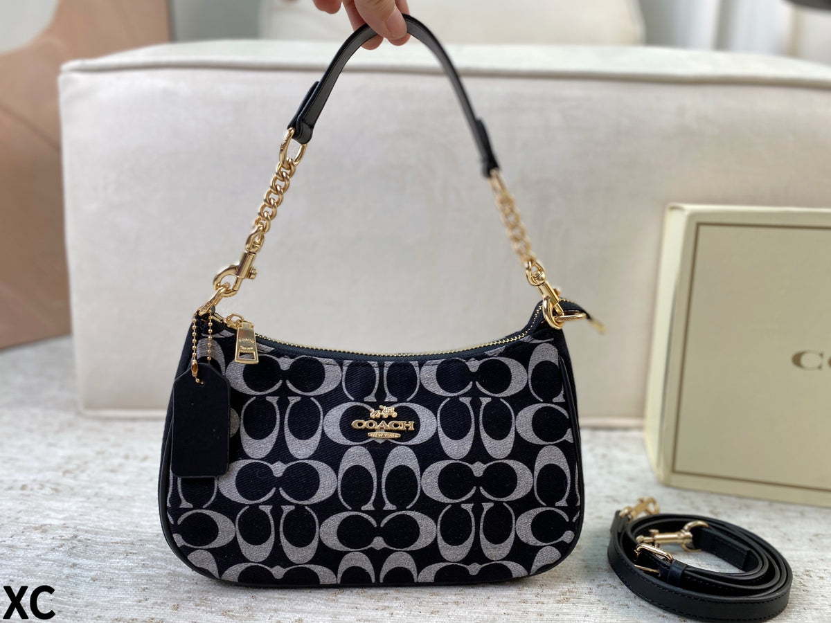 A Coach handbag with a black and gray signature monogram pattern, gold-tone hardware, and a black leather top handle. A hand is holding the handle, and a Coach box and a coiled strap are visible in the background on a light surface.