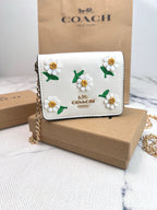 A Coach crossbody wallet in white leather with embroidered daisy flowers, displayed on a brown cardboard box with a Coach bag in the background.