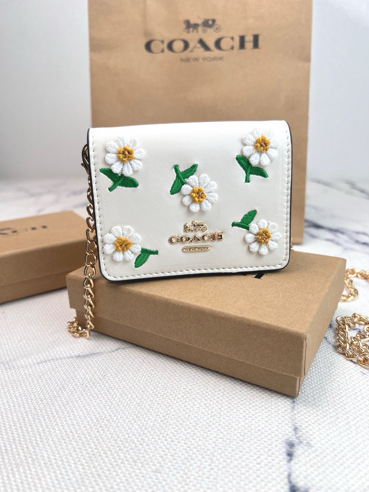 A Coach crossbody wallet in white leather with embroidered daisy flowers, displayed on a brown cardboard box with a Coach bag in the background.