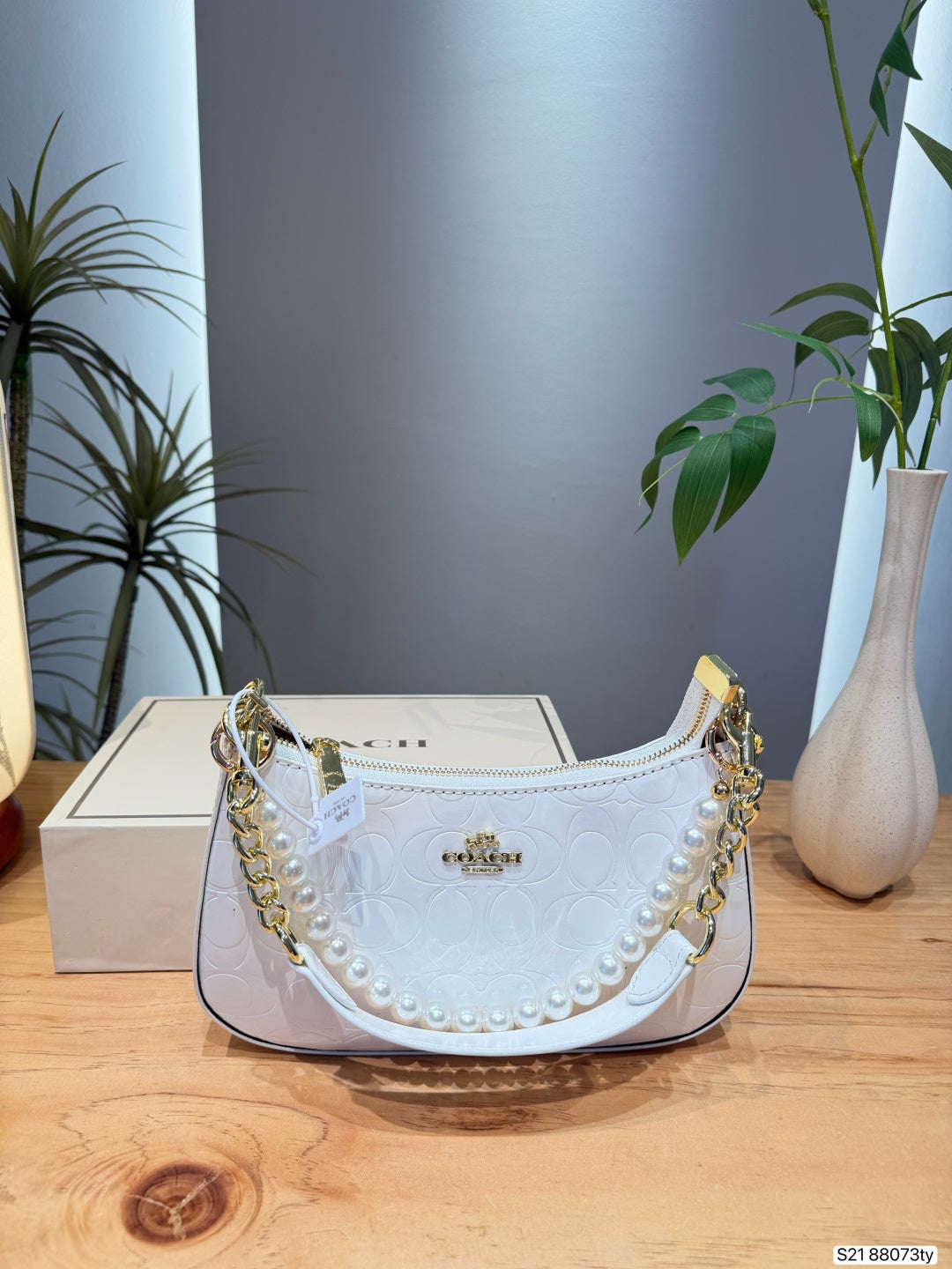 A white Coach crescent-shaped shoulder bag with a gold-tone chain strap, pearl trim, and a gold-tone logo clasp, displayed on a wooden table with its box and a vase in the background.