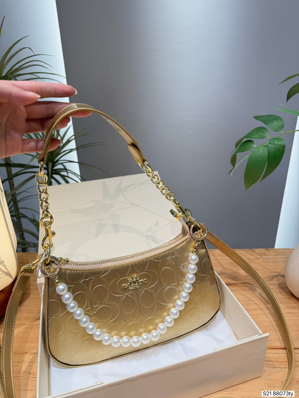 A gold-tone Coach handbag with a signature monogram pattern, a gold chain handle, and a decorative white pearl strand, resting on a white box with a hand holding the handle on the left.
