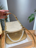 A gold-tone Coach handbag with a signature monogram pattern, a gold chain handle, and a decorative white pearl strand, resting on a white box with a hand holding the handle on the left.