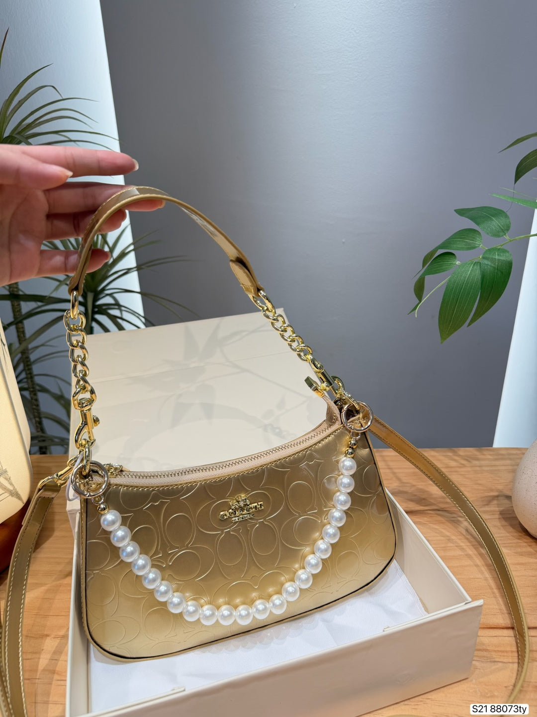 A gold-tone Coach handbag with a signature monogram pattern, a gold chain handle, and a decorative white pearl strand, resting on a white box with a hand holding the handle on the left.