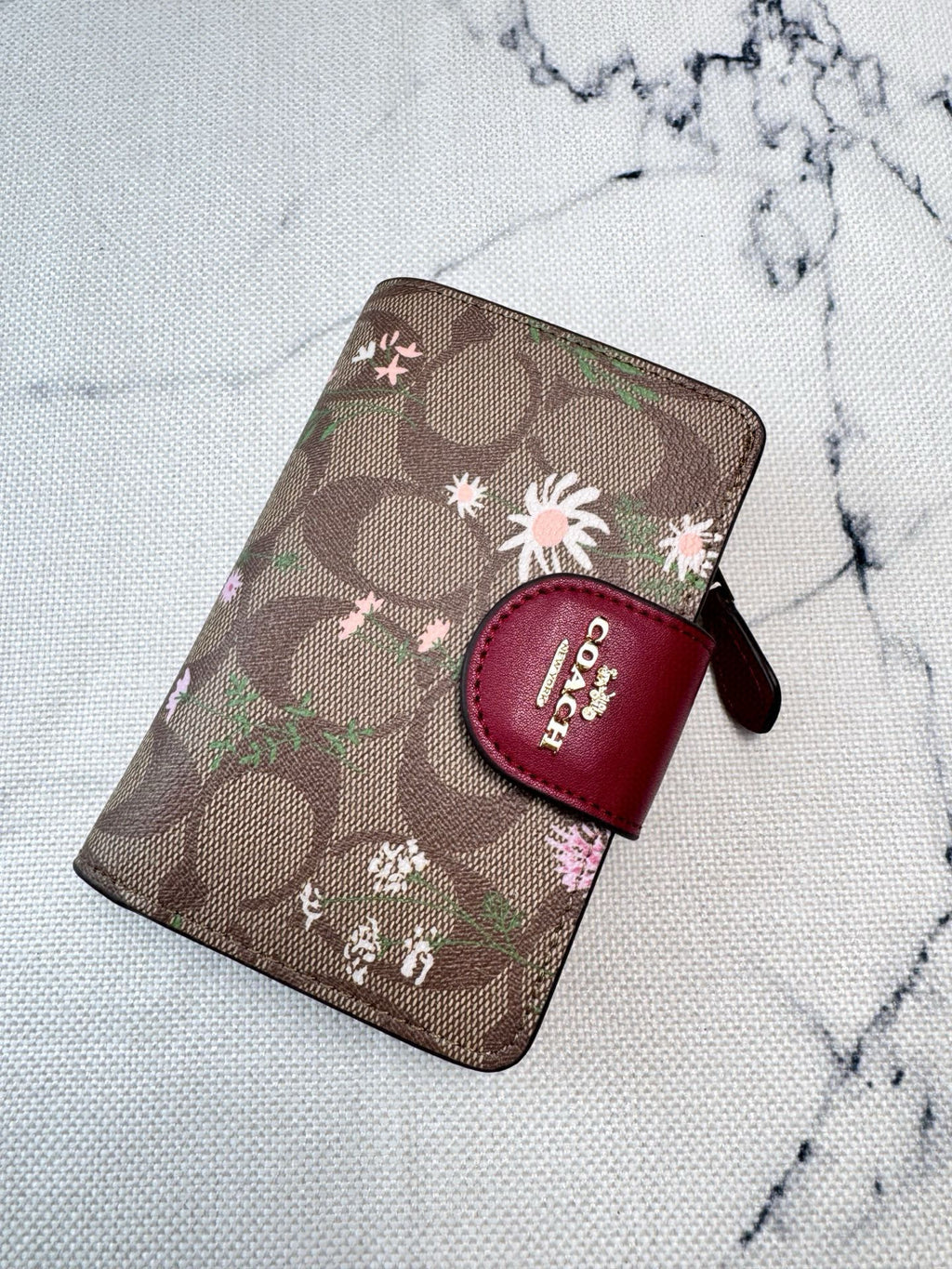 A Coach zip-around wallet in brown signature monogram canvas with pink and white floral prints, featuring a dark red leather logo tab on the front, photographed on a light marble-textured surface.