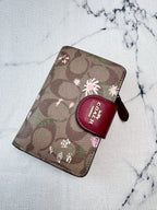 A Coach zip-around wallet in brown signature monogram canvas with pink and white floral prints, featuring a dark red leather logo tab on the front, photographed on a light marble-textured surface.