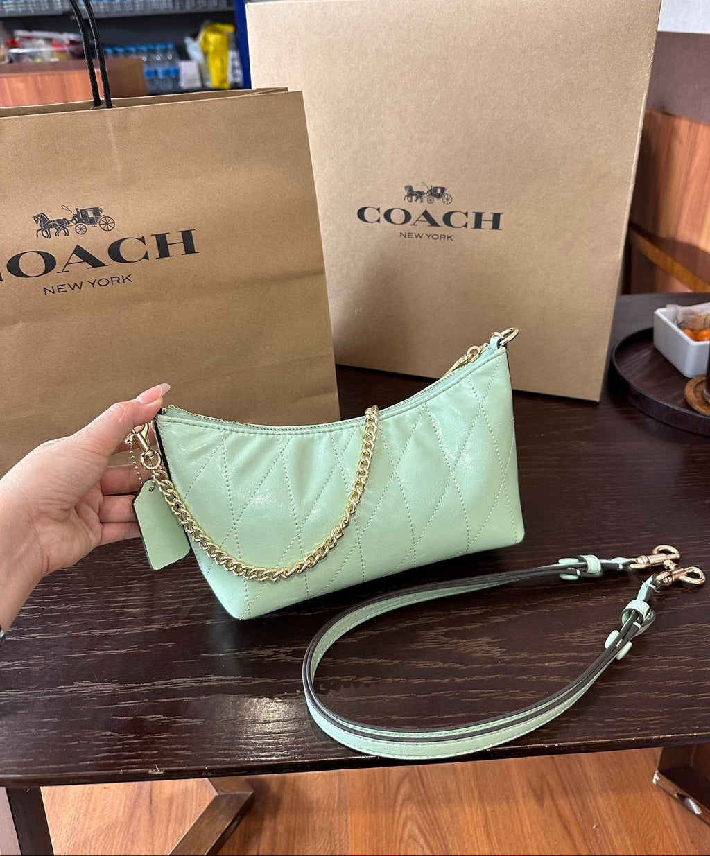 A light mint green Coach shoulder bag with quilted stitching and a gold chain link strap, held by a hand in front of two brown Coach New York shopping bags on a dark wooden table.