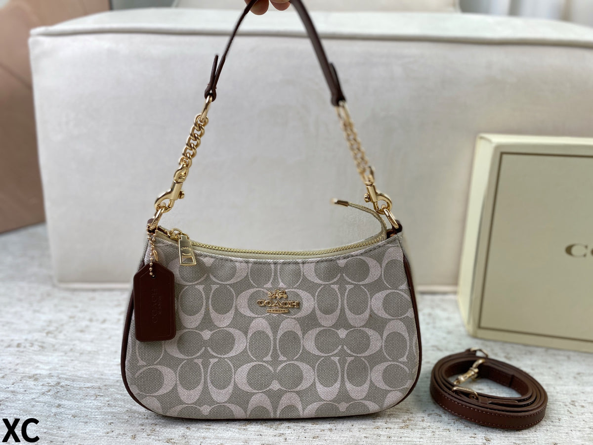 A Coach handbag in the signature monogram canvas, featuring a brown leather top handle, gold-tone chain strap, and a matching brown leather tag. A matching Coach box is visible in the background on a light-colored surface.
