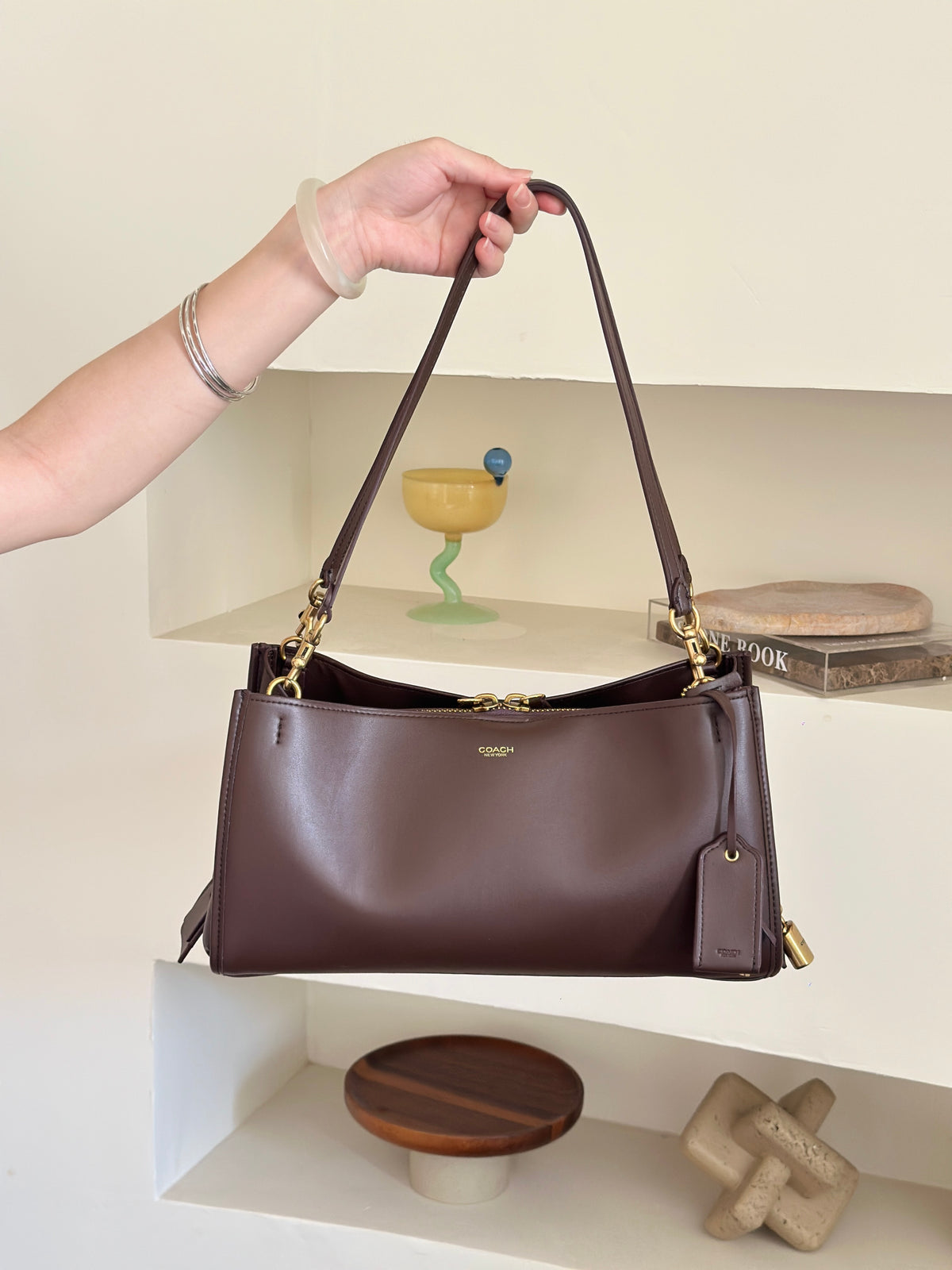A dark brown leather shoulder bag with gold-tone hardware and a small gold logo on the front, held by a person's hand against a cream-colored shelving background.