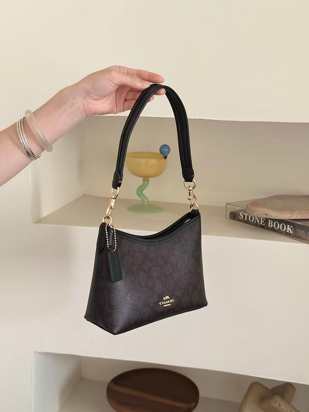 A hand holds a small dark brown monogram shoulder bag with gold-tone hardware and a top handle. The bag has a dark green leather tassel charm and a small gold logo on the front.