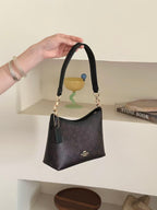 A hand holds a small dark brown monogram shoulder bag with gold-tone hardware and a top handle. The bag has a dark green leather tassel charm and a small gold logo on the front.