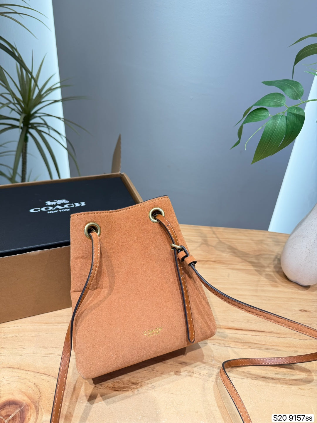 A Coach tan leather bucket bag with gold-tone hardware, displayed on a wooden table next to its black branded box.