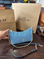 A blue Coach shoulder bag with gold-tone chain and strap hardware, held by a hand in front of two Coach paper shopping bags on a dark wooden table.