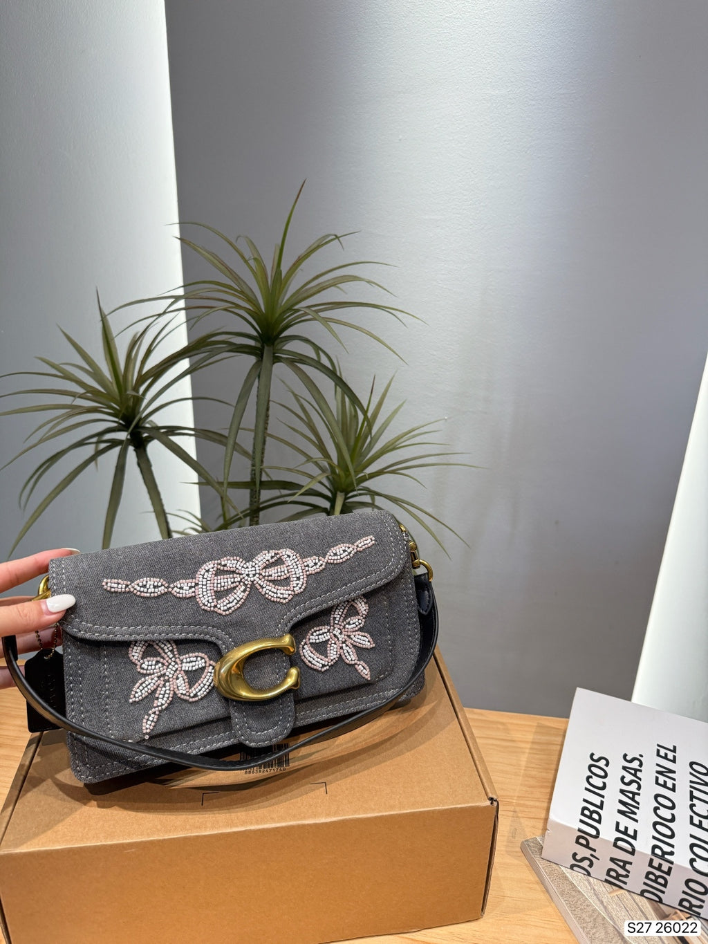 A gray suede shoulder bag with white embroidered bow and floral motifs and a gold-tone C-shaped clasp, held by a hand and resting on a brown cardboard box.