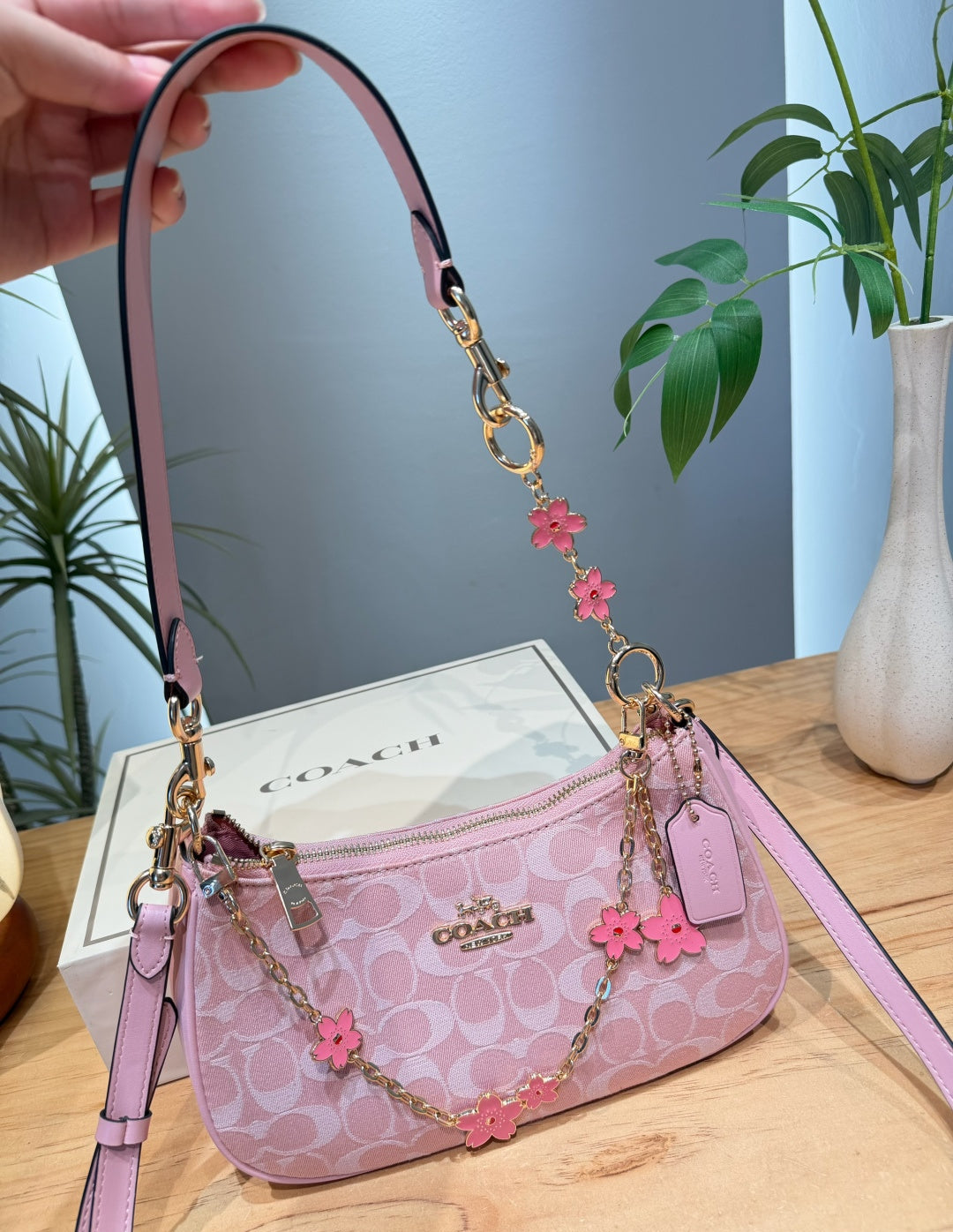 A pink Coach signature cante bag with a floral chain strap and pink flower charms, held by a hand in front of its white box on a wooden table.