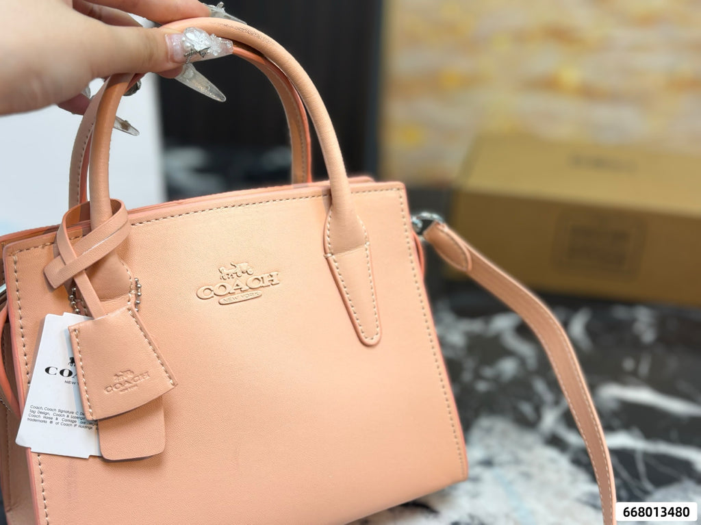 A peach-pink Coach handbag with a top handle, leather strap, and embossed logo, held by a hand with silver nail art. A white Cozzi tag is attached, and a cardboard box is visible in the blurred background.