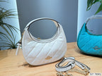 A white quilted Coach hobo handbag with gold hardware and a matching shoulder strap lies on a wooden table, with a turquoise Coach bag partially visible in the background and a blue wall behind it.