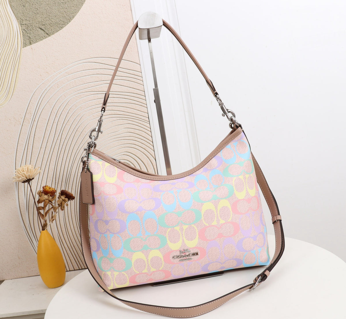 A pastel multicolor Coach hobo shoulder bag with a signature monogram pattern, displayed on a white table against a modern beige and white background.