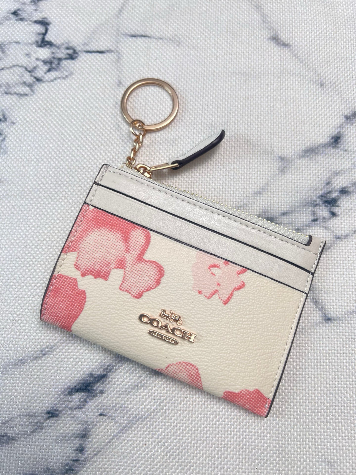 A Coach card holder keychain pouch with a cream and pink floral pattern, featuring a gold-tone key ring and a black leather pull tab, displayed on a white marble-patterned fabric background.