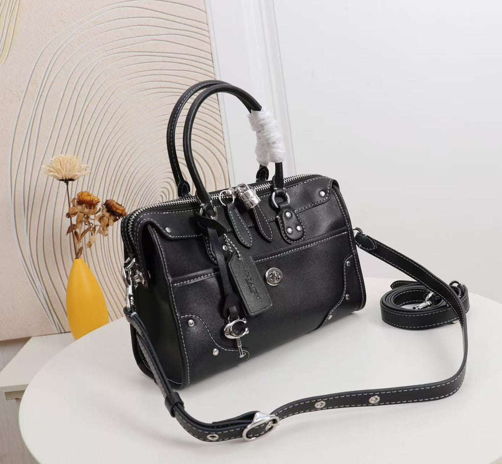 A black leather handbag with silver-tone hardware, dual top handles, a detachable shoulder strap, and a hanging keychain, displayed on a white round table with a decorative background.