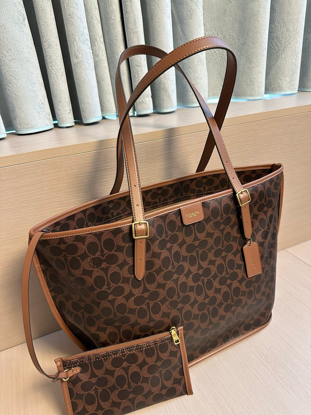 A brown and black Coach tote bag with tan leather handles and trim, shown alongside a matching zip pouch on a light wood surface in front of gray curtains.