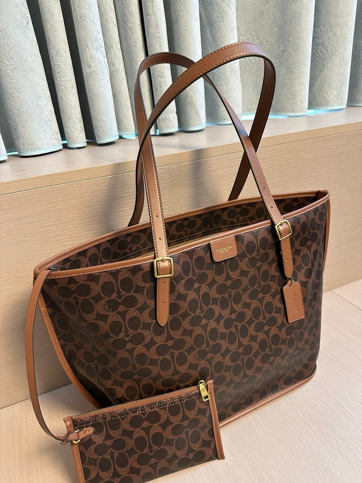 A brown and black Coach tote bag with tan leather handles and trim, shown alongside a matching zip pouch on a light wood surface in front of gray curtains.