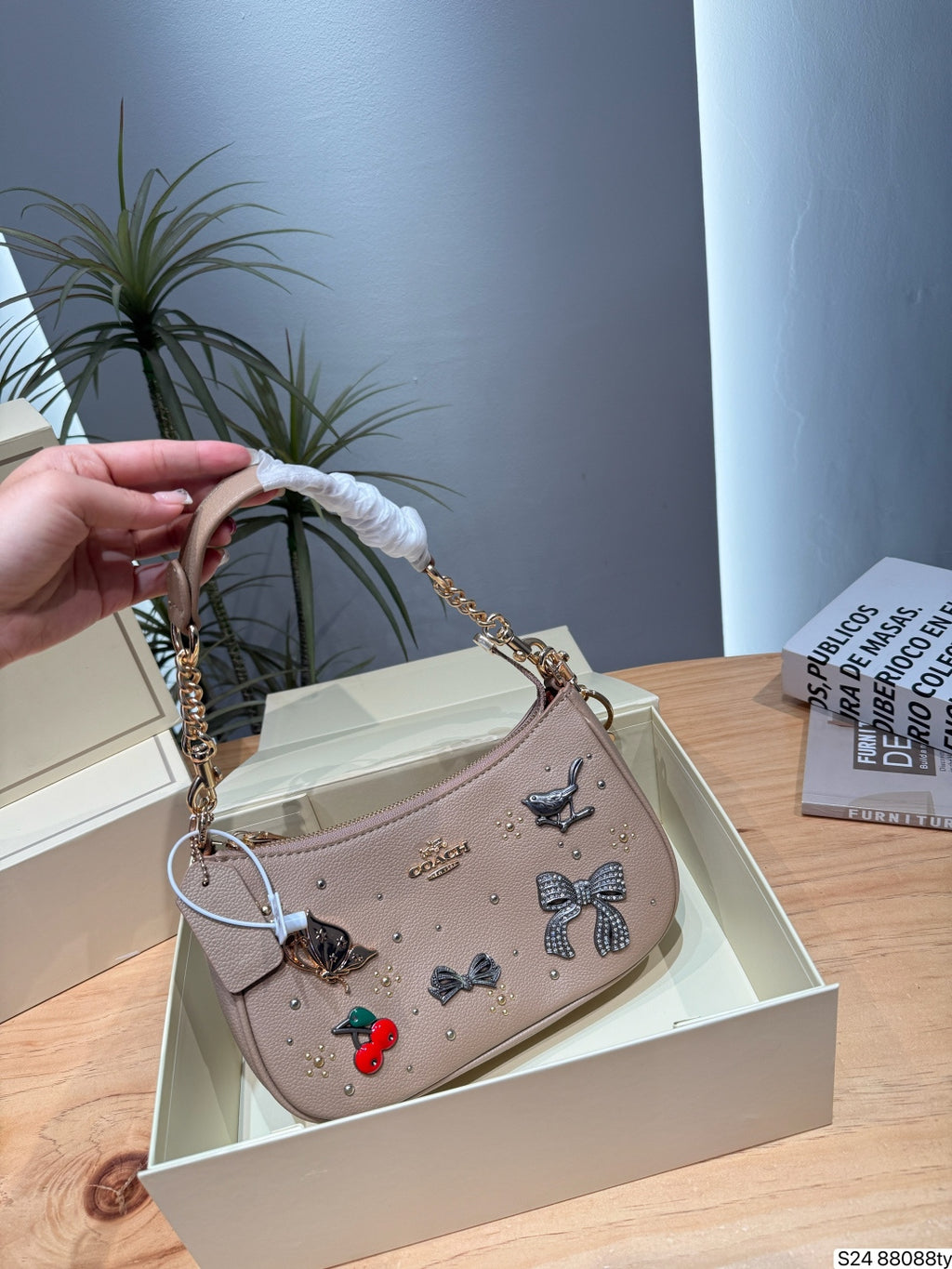 A beige Coach shoulder bag with decorative embroidered bows, butterflies, and cherries, held by a hand over an open cream-colored box on a wooden table.