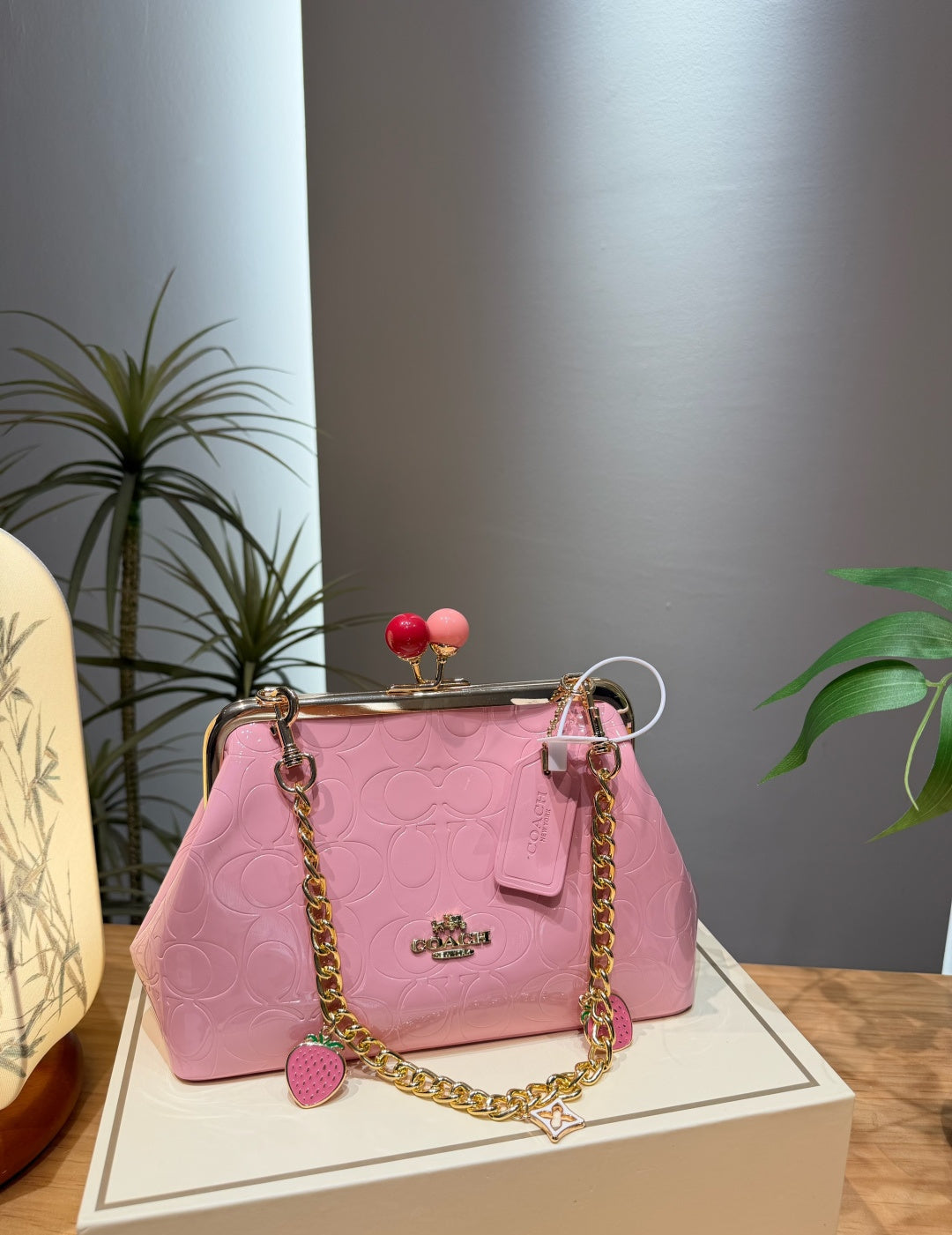 A pink embossed leather handbag with a gold-tone chain strap, kiss-lock closure, and strawberry charms, displayed on a white box in an indoor setting.