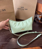 A Coach shoulder bag in pale mint green quilted leather with a gold chain strap, held by a hand in front of Coach shopping bags on a dark wooden table.