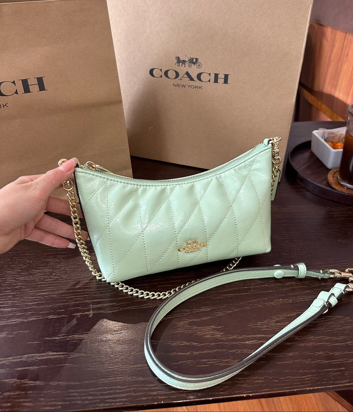 A Coach shoulder bag in pale mint green quilted leather with a gold chain strap, held by a hand in front of Coach shopping bags on a dark wooden table.