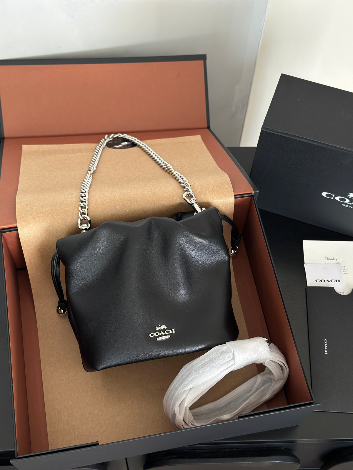 A black leather Coach handbag with a silver chain strap, presented inside its open black and brown box alongside a white protective wrap and a Coach box.