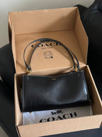 A black Coach handbag with gold-tone hardware resting inside an open brown cardboard Coach box, with the Coach logo visible on the inside lid and dust bag.