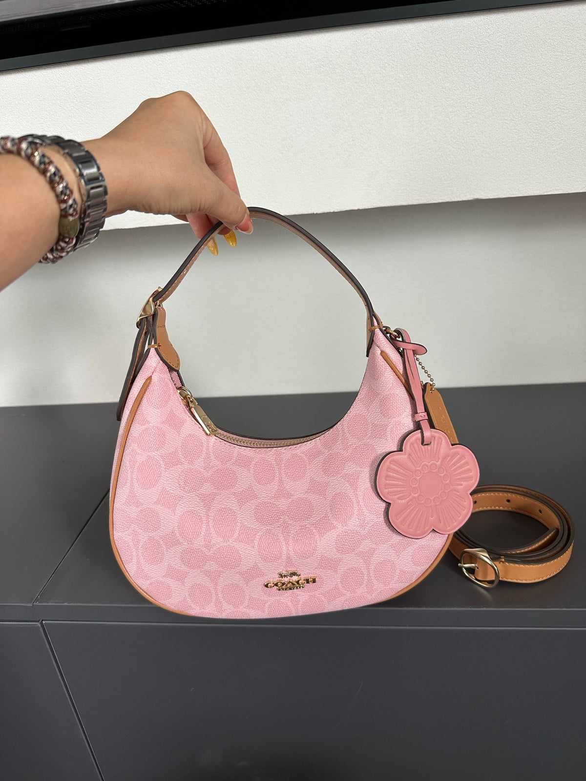 A pink Coach hobo shoulder bag with a signature monogram pattern, held by a hand. The bag features a pink flower-shaped charm, a gold-tone logo plate on the front, and a tan leather handle and strap.